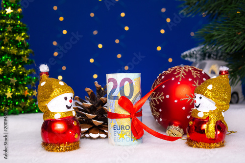 Between two snowmen is a euro banknote with a pine cone and a ball
