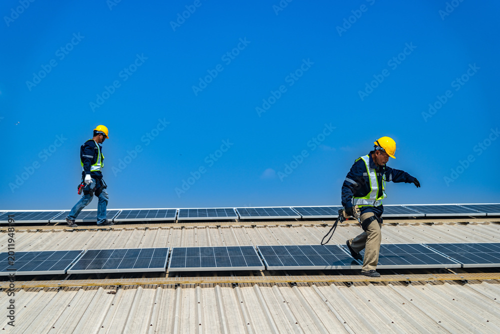 Fototapeta premium A professional technician in safety gear standing on a rooftop, preparing electrical wiring for a solar panel installation under a clear blue sky.