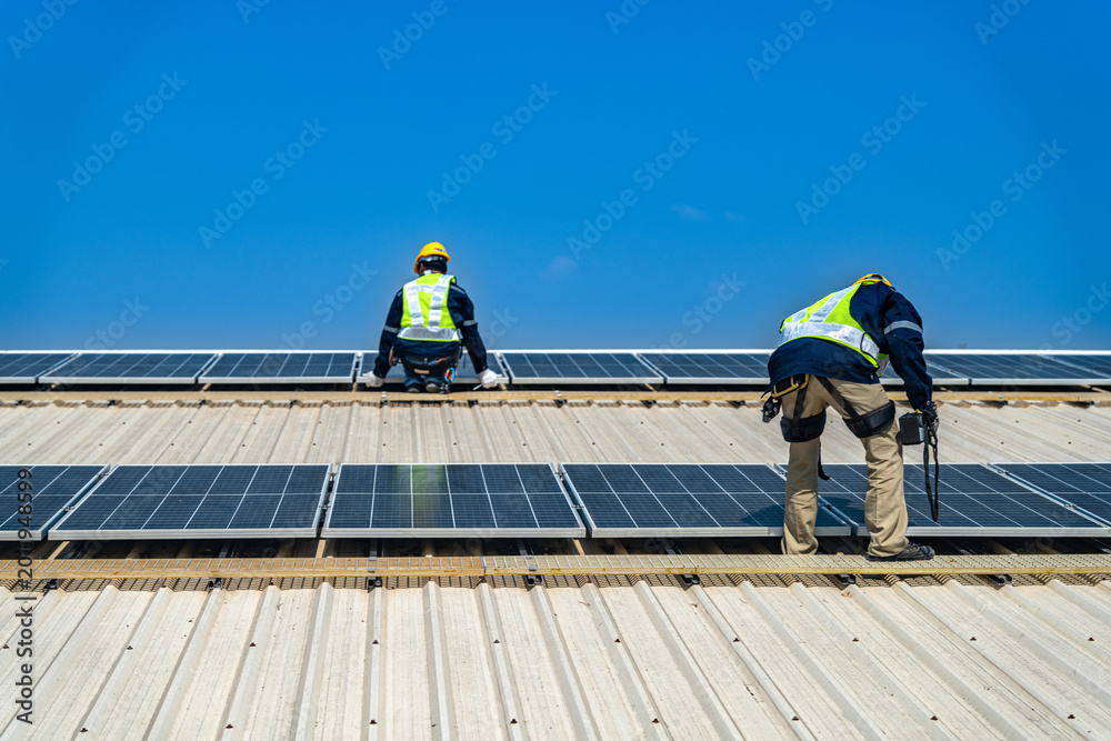 Fototapeta premium A professional technician in safety gear standing on a rooftop, preparing electrical wiring for a solar panel installation under a clear blue sky.