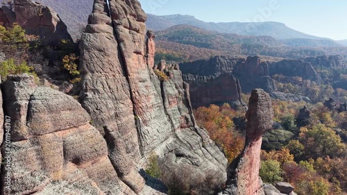 Close aerial pass along natural stone cliffs and a tall rock pillar above colorful woodland.