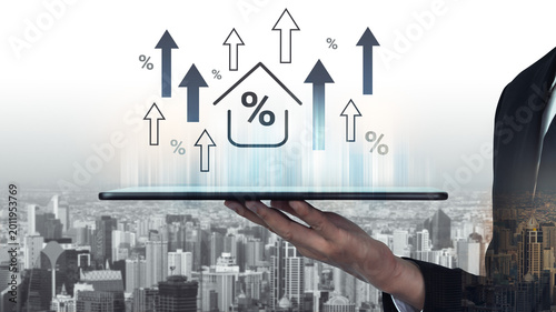 Photos A businessman presents a tablet displaying growth icons and house graphics over a cityscape, symbolizing real estate market trends and financial analytics