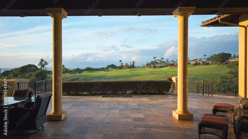 Fototapeta premium View from the terrace of a luxury house with large pillars that offers a view of the expansive green golf course and the sea in the distance at sunset