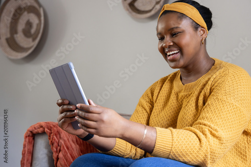 African American woman sitting at home holding gray tablet, wearing mustard sweater, orange throw