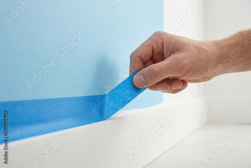 Close-up of a hand carefully peeling blue painter's tape from a freshly painted light blue wall.