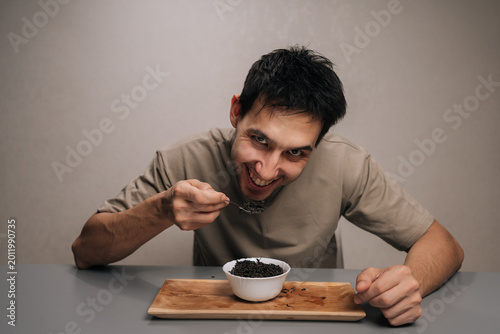 Foto Cheerful hungry young man with intense expression eating dry black tea leaves from bowl with a spoon, showing peculiar dietary habit or consumption of caffeine, smiling looking camera