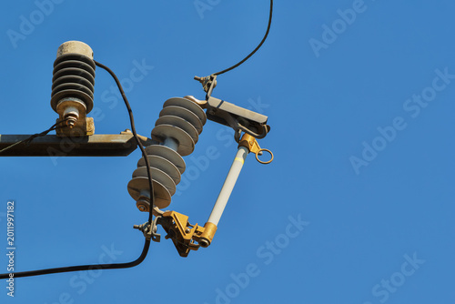 Electrical transformer fuse cutout on a utility pole with ceramic insulators and power lines against a clear sky, representing power distribution and utility infrastructure.