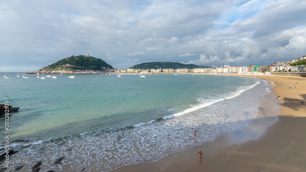 Obraz premium People walking near the shoreline of La Concha beach with calm waves and panoramic bay view in San Sebastian, Spain.