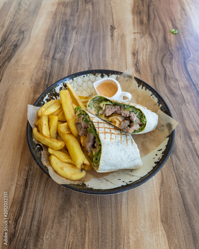 Close-up of a grilled beef wrap sandwich cut in half, served with crispy French fries and dipping sauce on a rustic ceramic plate. Casual fast food meal with natural lighting and wooden table backgrou