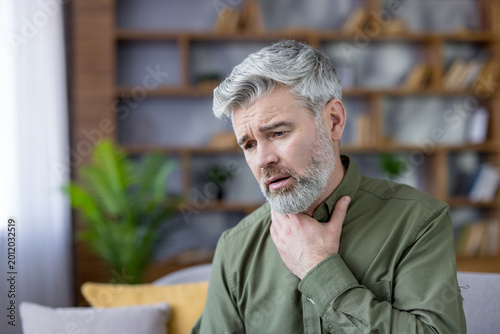Mature man touching his neck, experiencing pain and discomfort from a sore throat or difficulty swallowing, indicating a health problem or illness while indoors