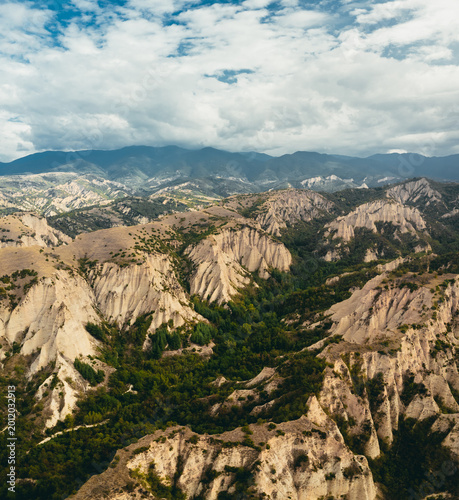 High-resolution aerial view of rugged sandstone canyon formations surrounded by dense green forest and rolling hills, with a distant mountain range beneath a dramatic cloudy sky. Rich natural textures