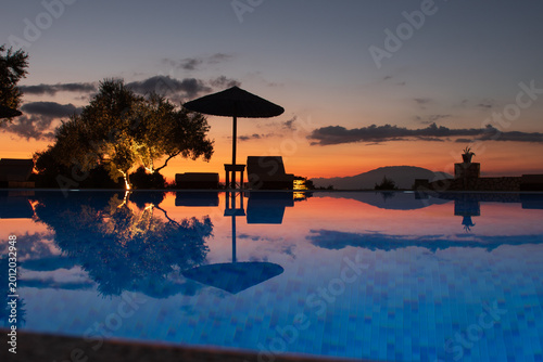 Valokuva Tranquil resort infinity pool reflecting dramatic sunset sky, with parasol, sunbeds, and warm evening light in a tropical holiday setting