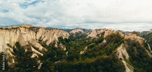 Wide panoramic drone view of eroded sandstone cliffs and rolling green valley with dense vegetation under a soft cloudy sky. Natural textures and unique geological formations create a dramatic and vis