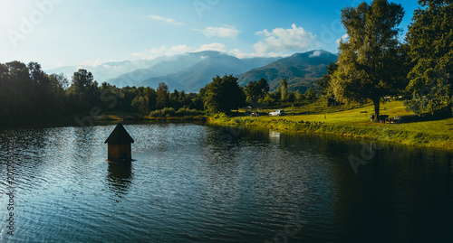 Peaceful landscape of a calm lake surrounded by green fields and trees with mountains in the background. A small wooden structure emerges from the water, illuminated by warm golden hour sunlight.
