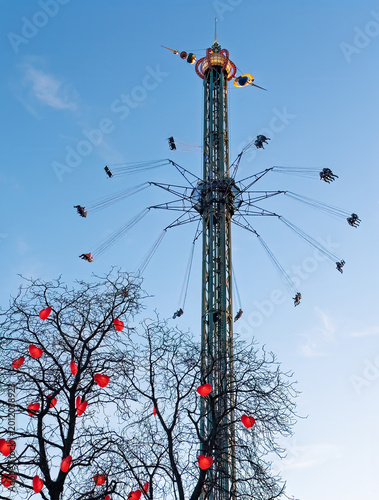 Copenhagen, Denmark - December 31, 2025: Tivoli Gardens. Flying in circles high above the ground against blue cloudscape, with red hearts in trees at bottom
