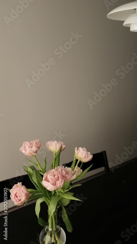 Modern, minimalist dining area and kitchen in a contemporary apartment. Sleek dark wooden dining table set with a glass vase of pink tulips, surrounded by designer chairs