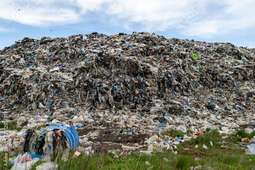 A large waste dump shows a tall heap of trash containing plastic, textiles, and other waste located in an open area