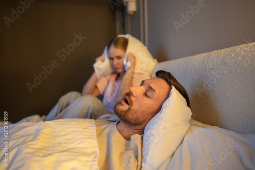 Φωτογραφία Woman stressed by loud annoying sound of husband snoring, sitting on bed covering ears with pillow, exhausted female with sleep deprivation and insomnia suffering from partner snoring at night