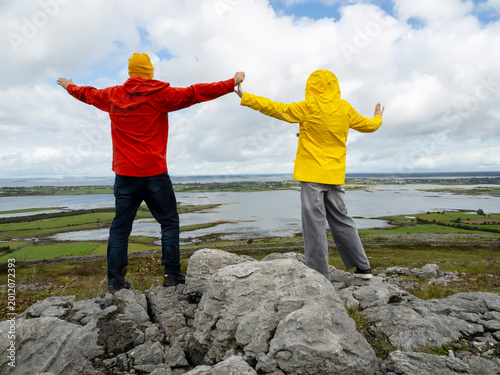 Father in red coat and daughter in yellow travel jacket are on top of a rough stone in a mountains, stunning nature scene with ocean and cloudy sky in the background. Divorced family concept. Travel.