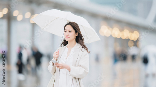 雨の街で白い傘を差しスマホを見ながら歩く若い女性
