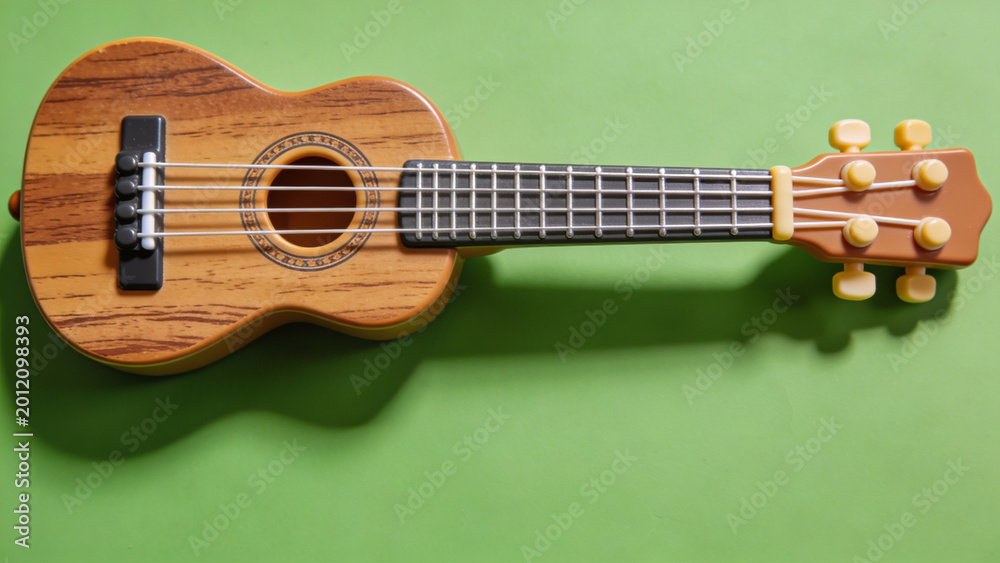 Fototapeta premium A wooden toy ukulele with four strings rests on a plain green surface.