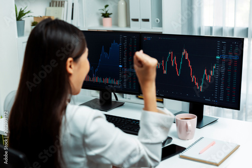 Schilderij op canvas Successful young Asian businesswoman earning highest profit with fist up in exchange stock market on dynamic business graph on pc screens at modern office, analyzing financial technology