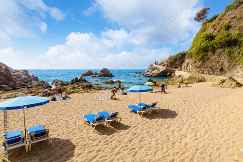 Canvas Print Sunbathers enjoy a sunny day along sandy Sa Caravera beach and rocky cove at the seaside resort town of Lloret de Mar, Spain, on the Mediterranean coast of the Costa Brava