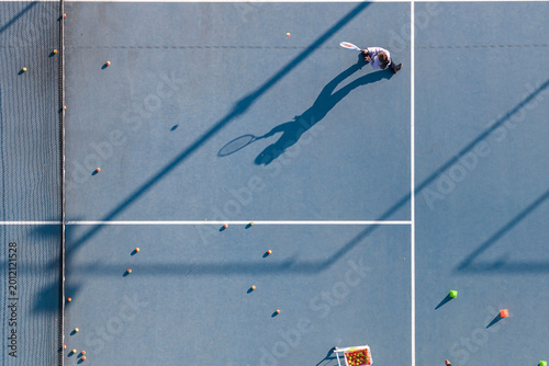 aerial view of a young boy in a yellow shirt playing tennis on a blue court. His long shadow captures his energetic swing. Multiple tennis balls are scattered.