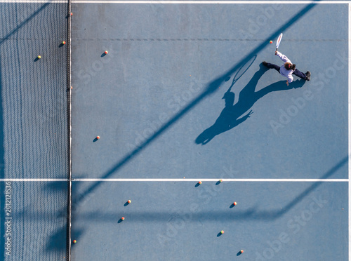 Quadro su tela Aerial view of a young girl playing tennis on a blue court, her long shadow naturally mirroring her dynamic swinging motion