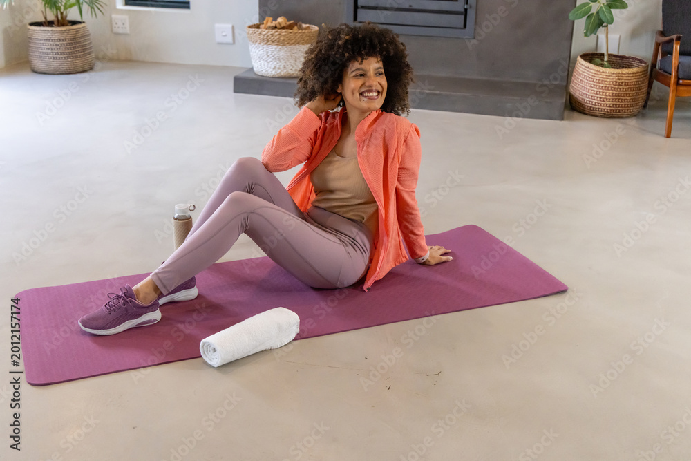 Obraz premium African American woman sitting on purple mat at home, wearing coral jacket with water bottle nearby