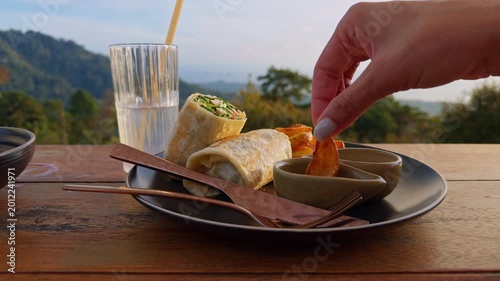 Hand Dipping Fried Snack Into Sauce On Plate With Wraps And Drink On Wooden Table, Tropical Mountain View Background, Golden Hour Light, Street Food Meal