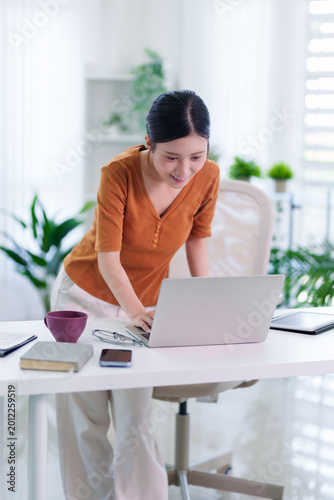 young Asian women working from home office using laptop computer hybrid remote work standing desk bright modern living room interior green plants technology