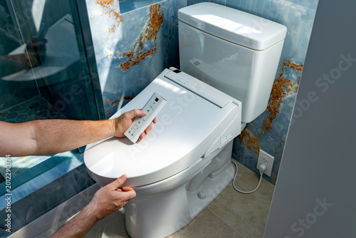 Man holding a remote control completing the installation of an electronic smart bidet toilet seat plugged into a wall outlet in a modern bathroom, USA