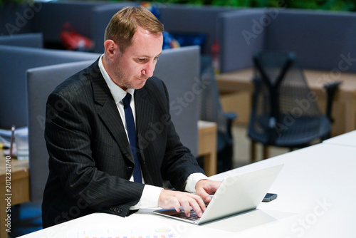 A professional senior executive or mature businessman in a black suit multitasking by using a laptop and smartphone. He is reviewing documents and communicating in a modern bright office environment. 