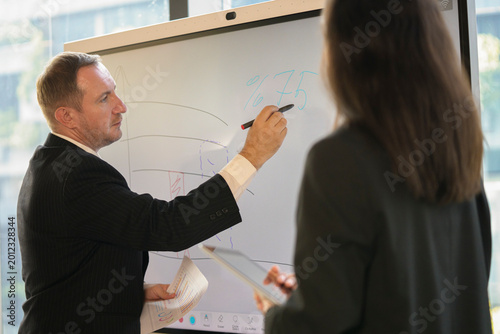 Senior executive mentoring young female colleague with whiteboard and laptop in office.