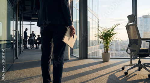 Tableau sur toile Professional businessman walking through a sunlit modern office hallway with a notebook in hand