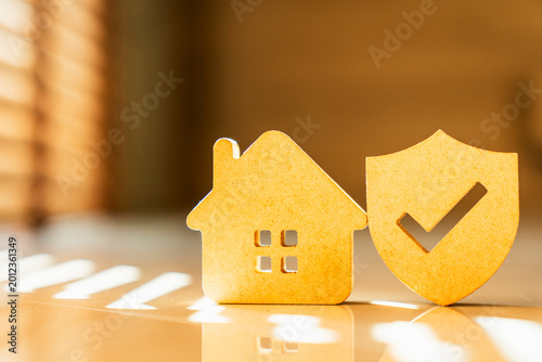 Wooden house model standing next to shield icon with check mark on sunlit floor, concept of home insurance, property protection, legal safety, residential security and asset guarantee.