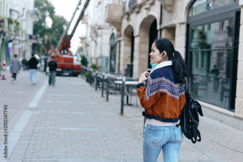 Konstfotografi Woman with backpack walks along a city sidewalk wearing a patterned jacket and jeans, glancing back as urban street scene unfolds with buildings and pedestrians