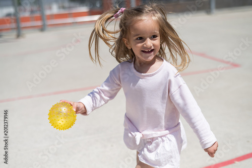 Happy preschool girl running across a sunny playground holding a yellow sensory ball, laughing and exploring outdoors in joyful, active play that promotes development and wellbeing