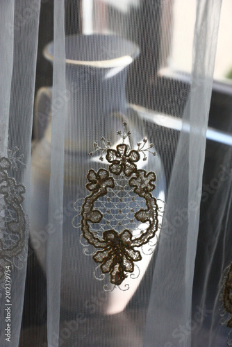 The vintage white jug stands on a window sill behind a transparent curtain. The original embroidery is on fabric.