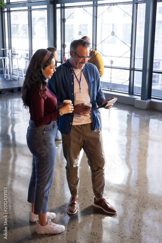 Diverse coworkers standing in modern lobby viewing tablet while holding coffee cups and lanyards
