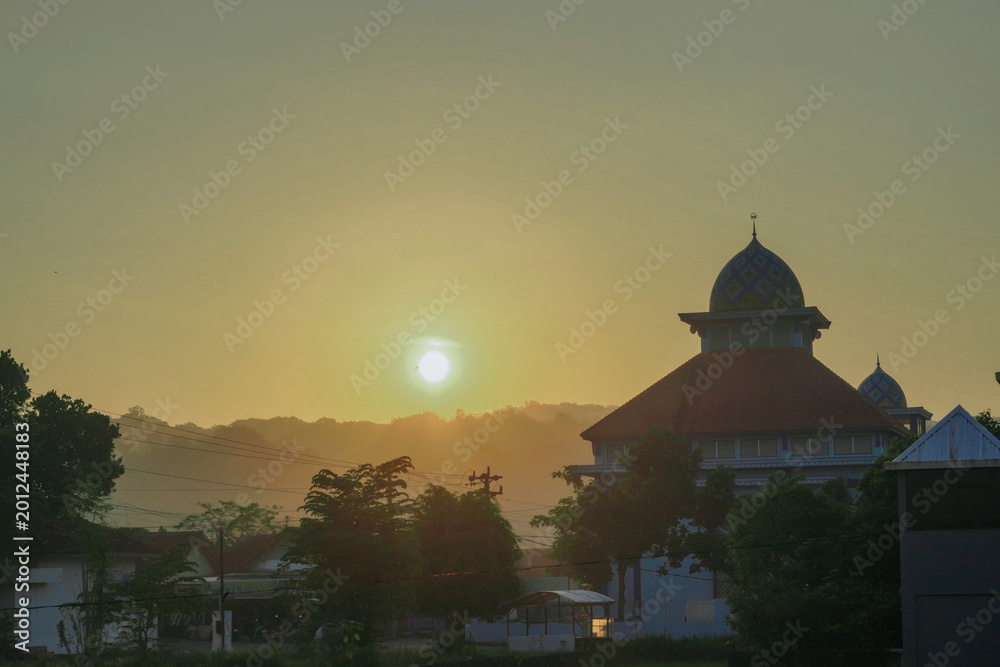 Obraz premium The serene view of the sunrise shows the silhouette of a mosque with a colorful dome standing majestically among the trees and residential areas.