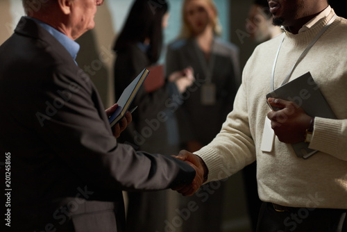 Close up of Black young adult man and senior Caucasian businessman shaking hands during networking event, holding tablet and badge. Ideal for partnership, leadership, hiring campaigns