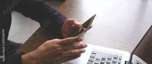 Close-up of hands using smartphone beside laptop on wooden desk, representing online communication, remote work, mobile technology, and digital lifestyle