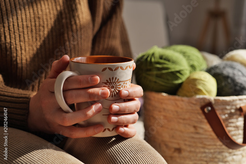 A person sits with a mug in their hands, surrounded by a basket filled with colorful yarn. The light from the window creates a warm atmosphere