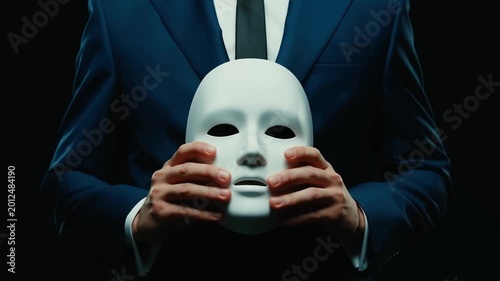 Man in suit holds white mask against dark background. Hands gently cradle mask, suggesting mystery or anonymity. Dark setting adds dramatic tone. Suitable for themes of secrecy, identity