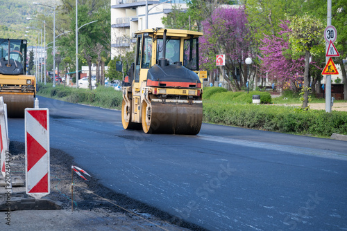 Canvas-taulu A yellow road roller compacts a fresh layer of smooth black asphalt on a city street during a road reconstruction project