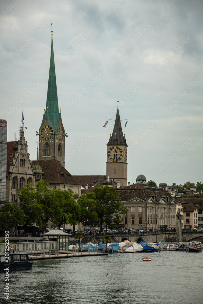 Fototapeta premium View of Fraumünster and St. Peter Church Towers over the Limmat River on a Cloudy Day in Zurich, Switzerland
