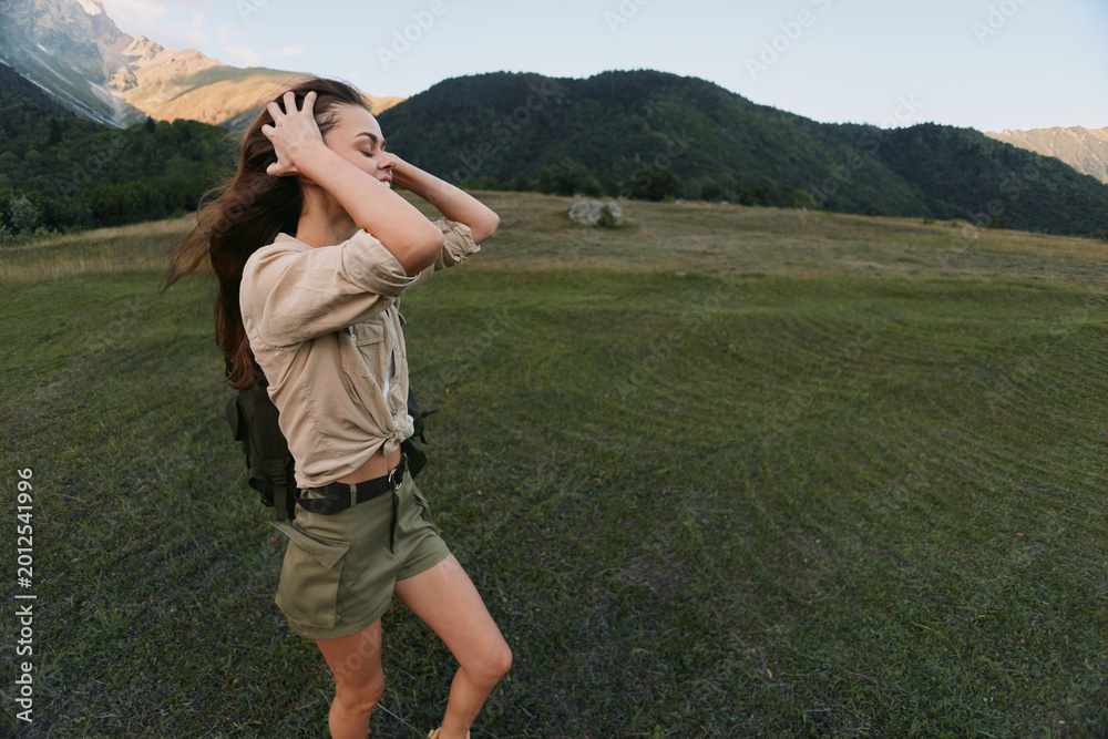 Naklejka premium Woman hiking in a grassy valley, adjusting hair, wearing beige shirt and shorts, backpack ready, surrounded by mountains and wide open sky, vibrant outdoor lifestyle moment