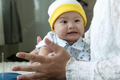 Bilde på lerret Close up of an unhappy Asian baby wearing a yellow beanie crying and looking upset while being held and comforted by mother's hands