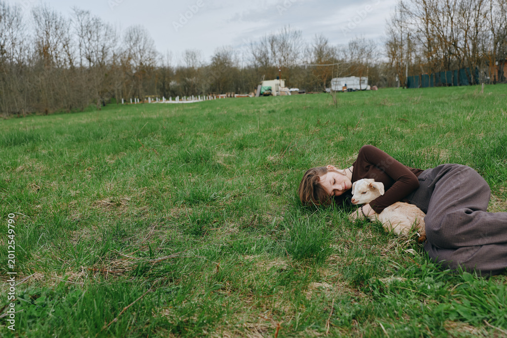 Naklejka premium A woman lies on the grass in a park, cuddling a dog as soft sunlight washes the scene, conveying relaxation, companionship, lifestyle, park, tranquil, candid moments outdoors.